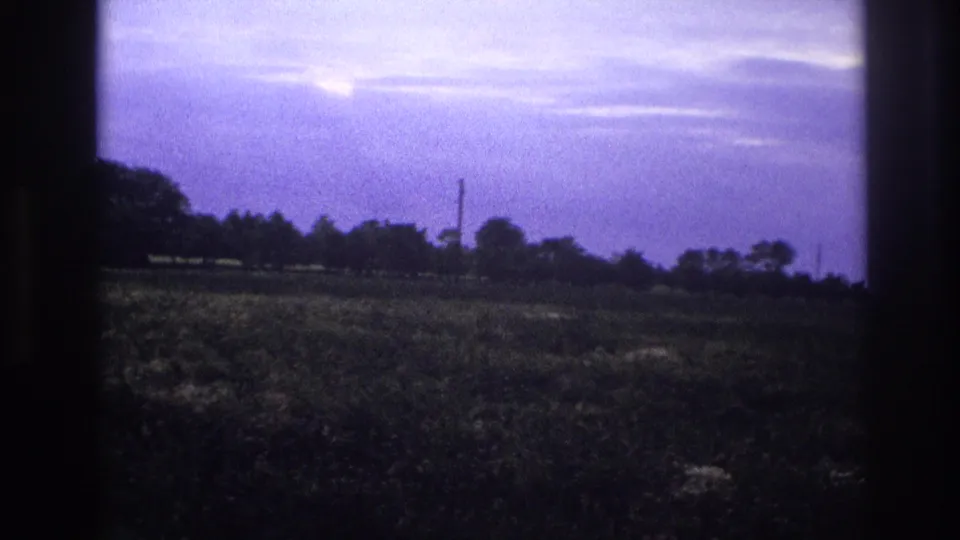 Continuation of 8mm vehicle-shot footage, rural France c.1967 — flat agricultural field, purple-cast sky, tree line and utility pole.