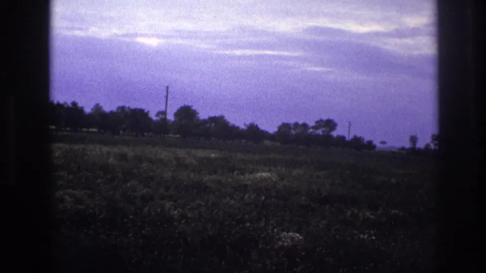 8mm home movie frame, rural France c.1967 — camera tilted to show expansive purple-cast sky over a low tree line and grassy field.