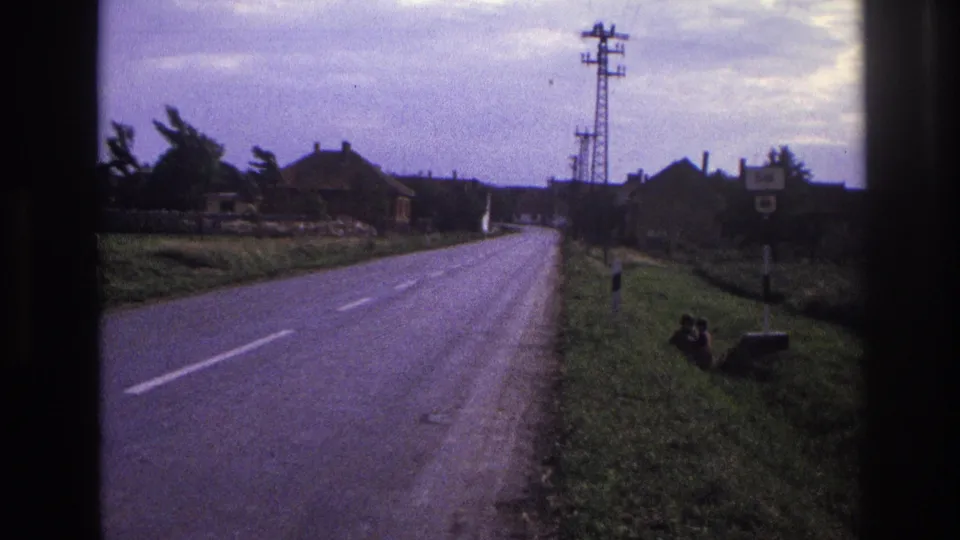 8mm vehicle-shot view down a French rural road, c.1967 — farmhouses left, electricity pylon right, dashed centre line, small figure seated on verge.