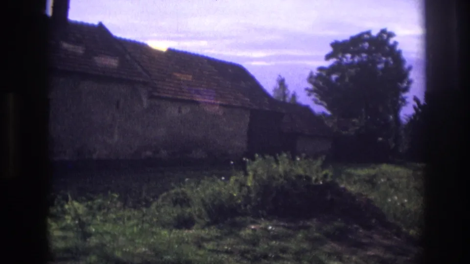 1967 France home-movie frame showing the side of a rustic house, tiled roof, rough wall surface, greenery, and nearby trees.
