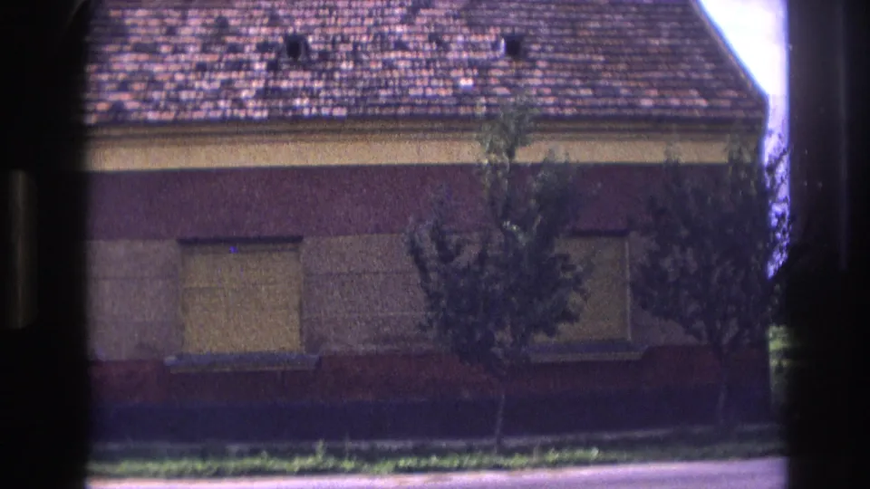 1967 France frame showing the front of a countryside house with shuttered windows, tiled roof, small tree, grass strip, and road edge.