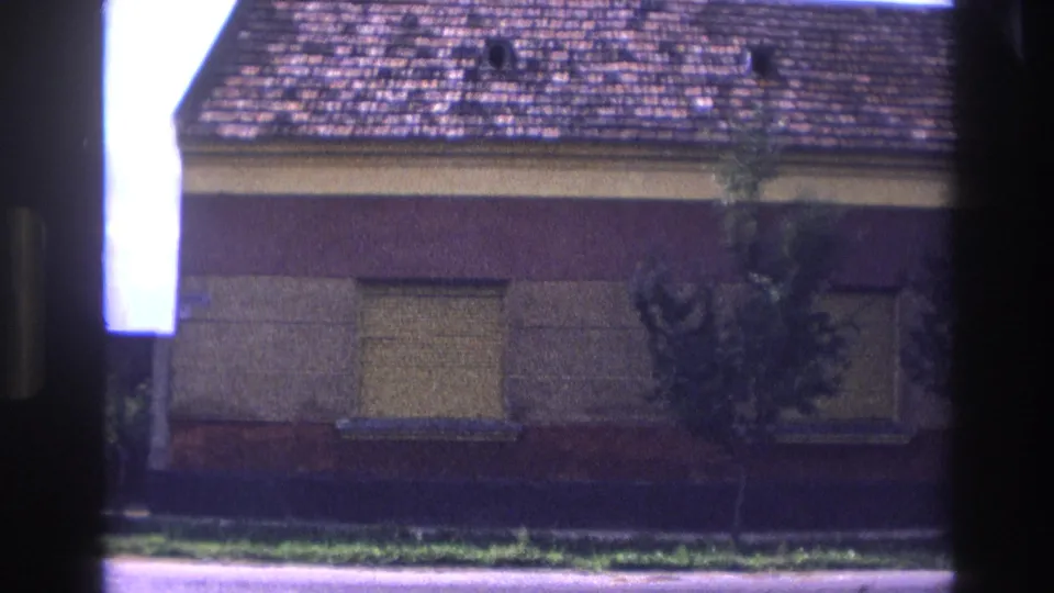 1967 France 8mm frame with a slightly tilted street view of the red and yellow house, boarded windows, roof tiles, and small trees.