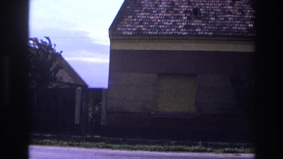 1967 France 8mm frame showing part of the house, one boarded window, a tiled roof, fence line, street edge, and adjacent roof shape.