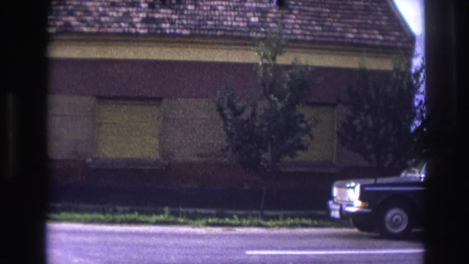 A dark classic sedan begins to pass the shuttered house on a residential street in France, 1967.