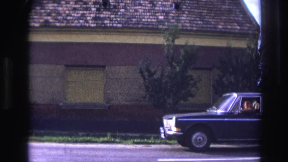 A dark sedan passes the shuttered house, with occupants faintly visible through the side window.