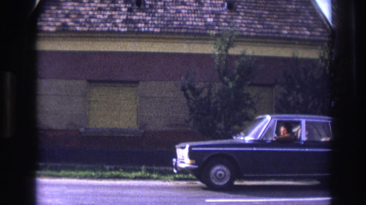 1967 France 8mm frame showing a dark blue sedan with visible occupants passing a shuttered building and tiled roof.
