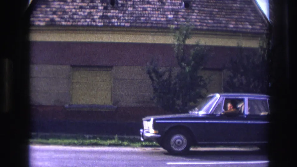 1967 France 8mm frame showing a dark blue sedan with visible occupants passing a shuttered building and tiled roof.