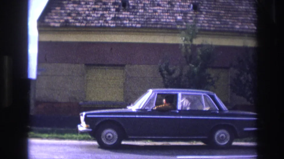 1967 France home-movie frame of a dark blue sedan moving along a residential street with occupants visible inside.
