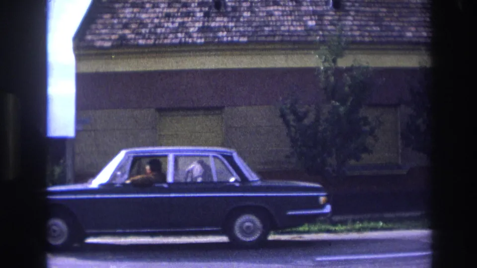 1967 France 8mm frame showing the side of a dark blue sedan, with occupants visible as it moves past a shuttered house facade.