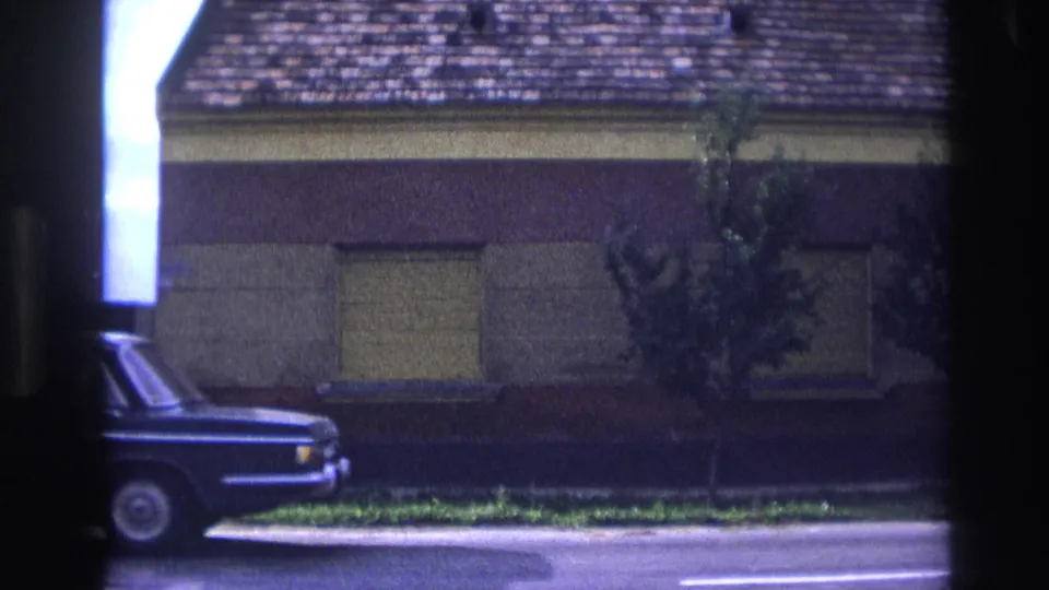 1967 France home-movie frame with the sedan partly exiting the shot along a residential street with shuttered windows behind it.