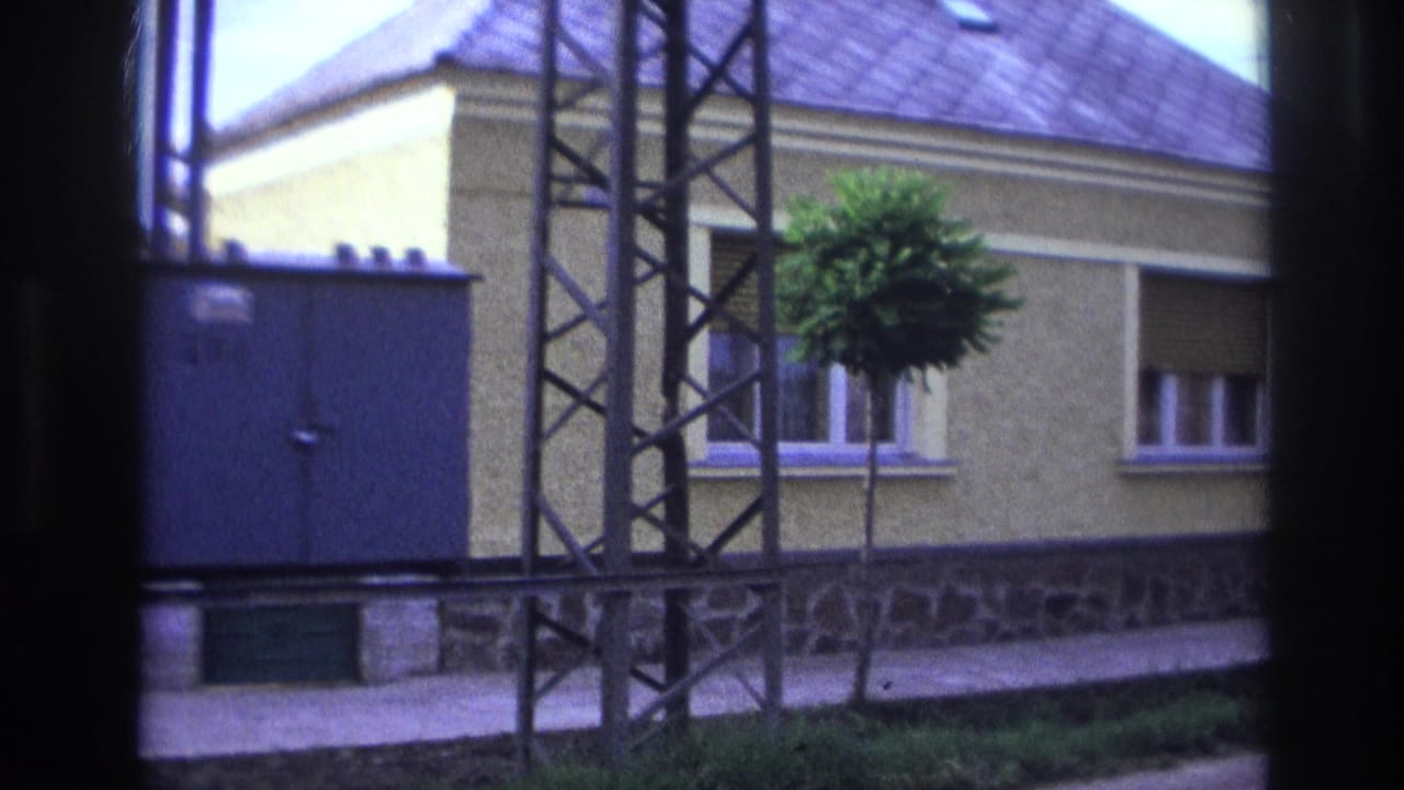 1967 France street view with a metal electrical structure, blue utility box, yellow house, stone foundation, and small sidewalk tree.