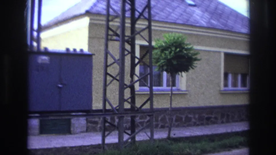 1967 France street view with a metal electrical structure, blue utility box, yellow house, stone foundation, and small sidewalk tree.