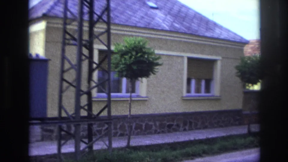 1967 France 8mm frame with a yellow stucco house, metal tower, shuttered windows, sidewalk, grass verge, and trimmed street trees.