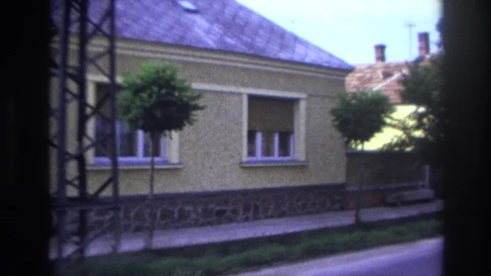 1967 France residential street frame showing a yellow house, closed shutters, tiled roof, stone base, sidewalk, and small trees.