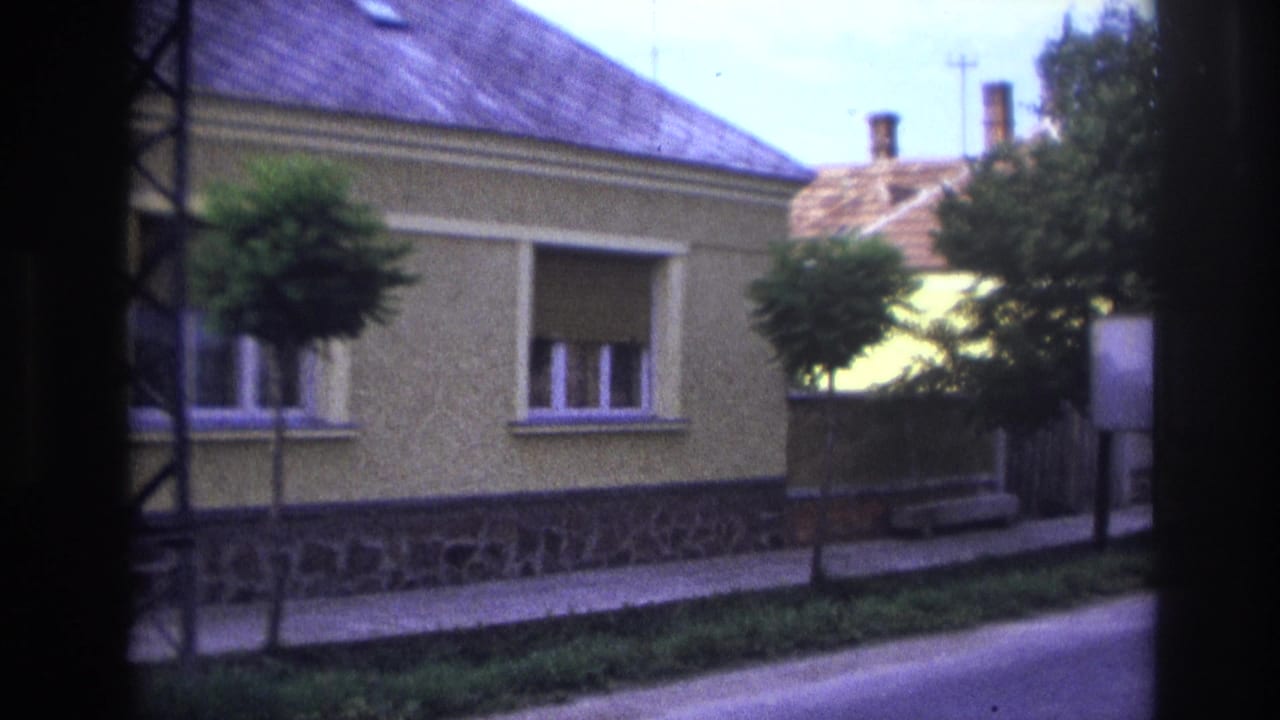 1967 France home-movie frame showing a yellow stucco house, tiled roof, sidewalk, small trees, and roadside verge.