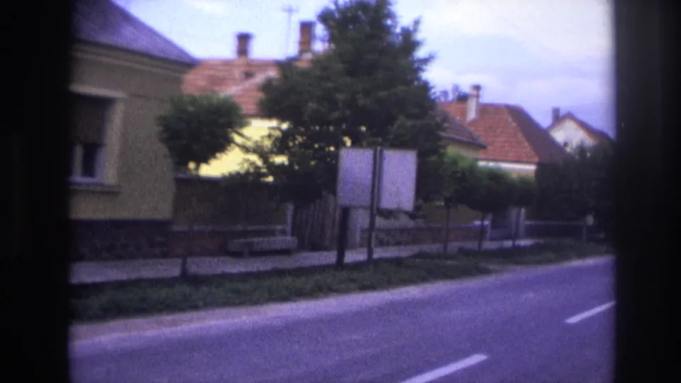 1967 France home-movie frame showing a paved road, sidewalk, sign boards, fences, trees, and low houses with tiled roofs.