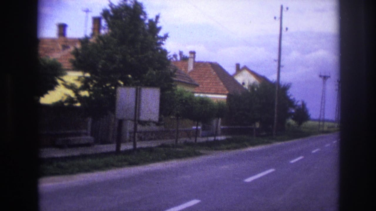 1967 France home-movie frame showing a quiet road, red-roofed houses, trees, roadside signs, and utility poles.