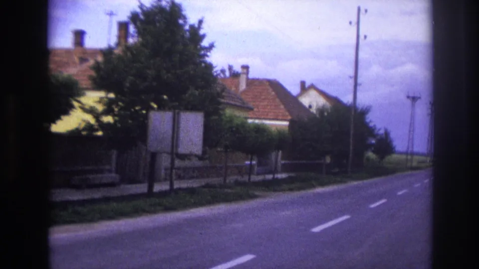 1967 France home-movie frame showing a quiet road, red-roofed houses, trees, roadside signs, and utility poles.