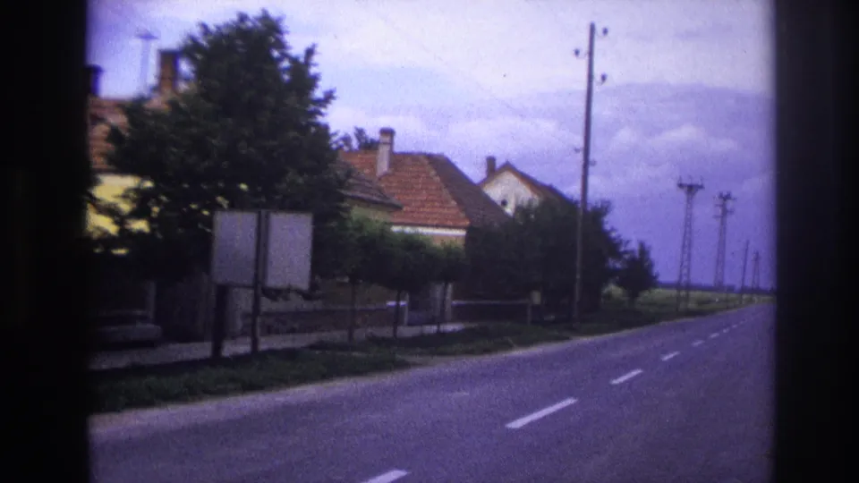 1967 France roadside view with red-roofed houses, hedges, utility poles, and an empty paved road.