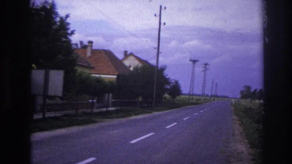 1967 France travel frame showing a quiet road receding past houses, trees, grass, and roadside utility poles.