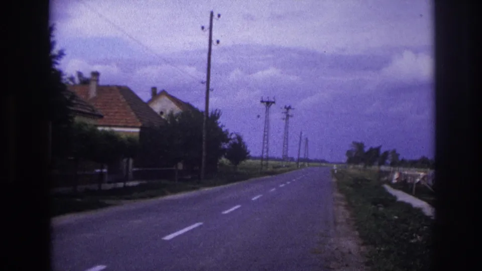 1967 France home-movie view of a rural street with houses, trees, utility poles, and grassy roadside edges.