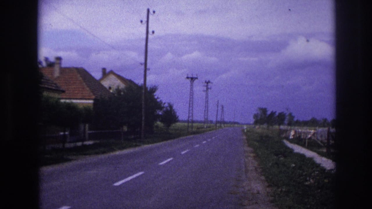 1967 France home-movie frame showing a rural road lined with utility poles and low houses under a cloudy sky.