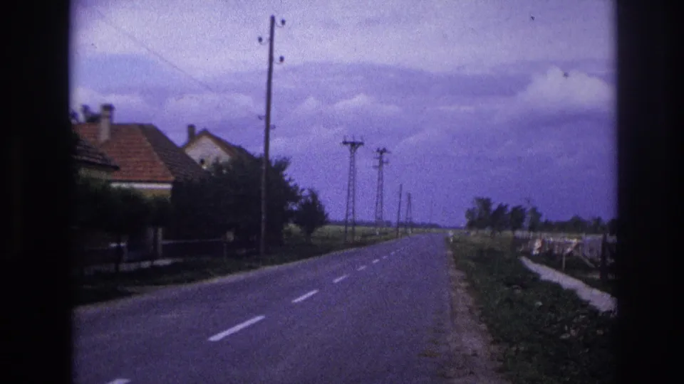 1967 France home-movie frame showing a rural road lined with utility poles and low houses under a cloudy sky.