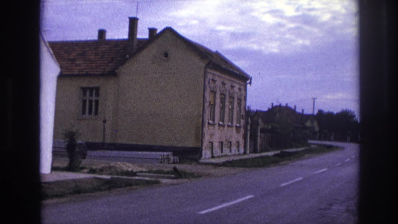 Pale stucco house beside a curving road with tile roof, chimneys, and cloudy sky.