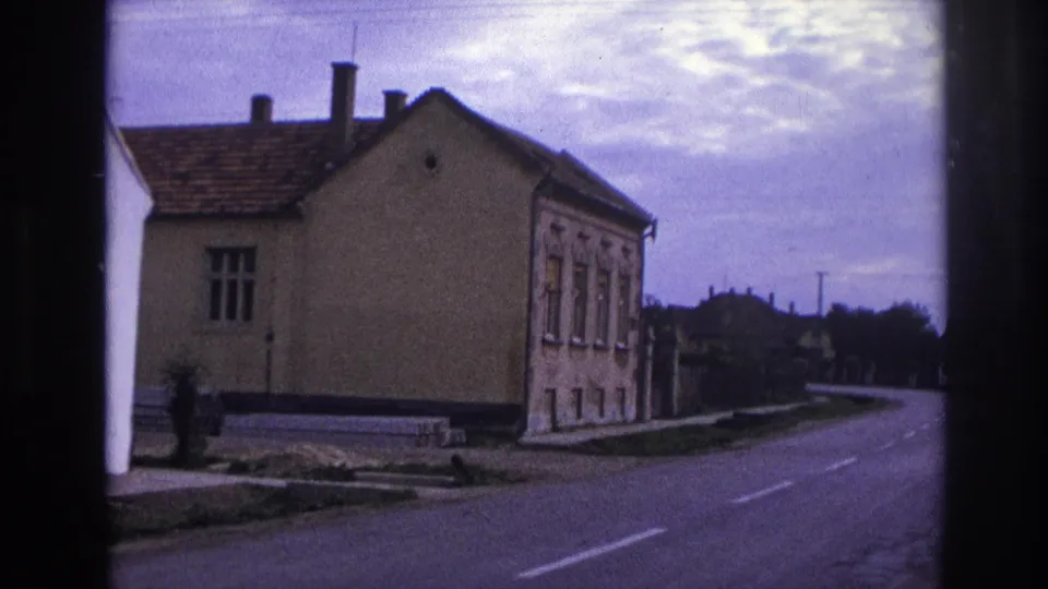 1967 France frame showing a stucco house beside a curving rural road with lane markings and distant rooftops.