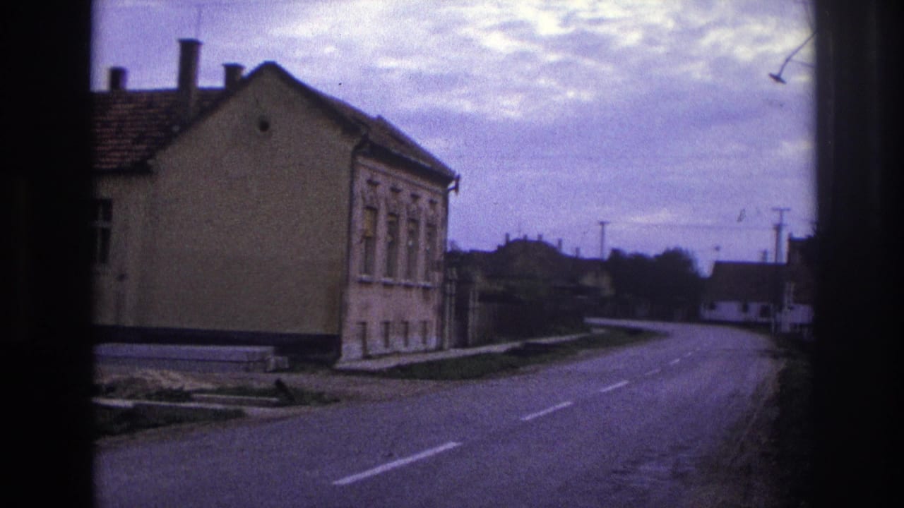 1967 French home-movie frame showing a quiet paved road curving past a modest two-story building with tiled roof and chimneys.