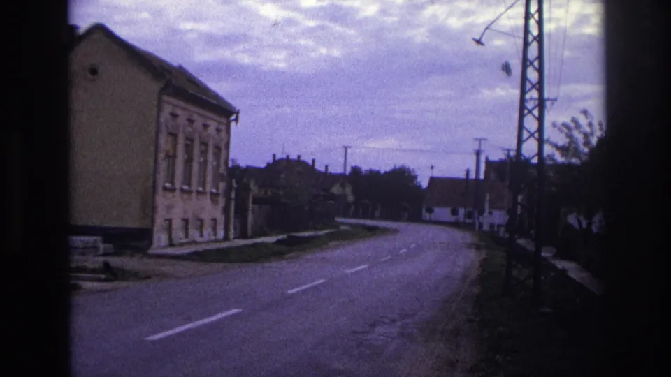 1967 French town road seen from a moving vehicle, with houses, overhead wires, roadside vegetation, and a lattice tower at right.