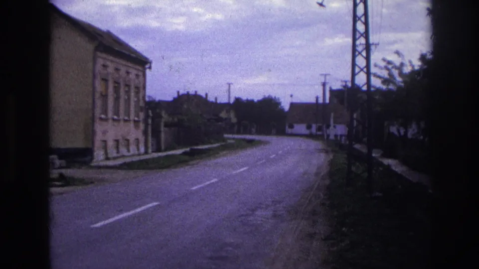 A moving-vehicle view of a French town road in 1967, with a two-story building, road markings, overhead lines, and a utility tower.