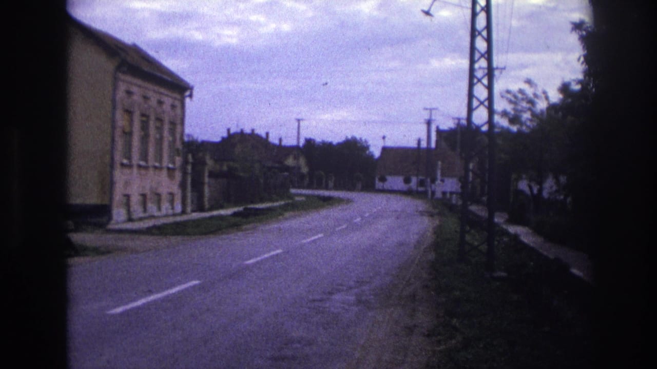 1967 France 8mm frame showing a quiet rural road with modest buildings, roadside greenery, utility poles, and an overcast sky.