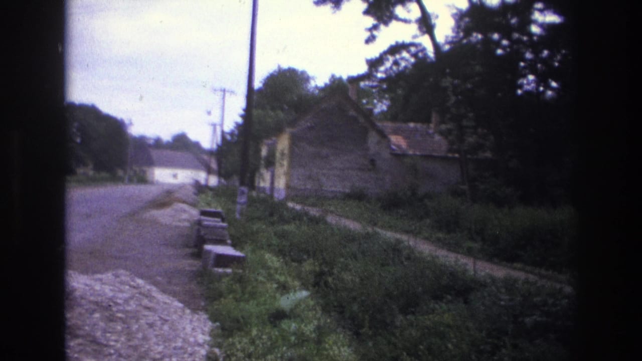 Roadside view with a small building, trees, grass, utility poles, and benches or low stone blocks.