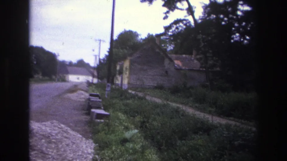 1967 France 8mm frame looking along a quiet roadside with a small stone building, trees, grass, utility poles, and low blocks near the verge.