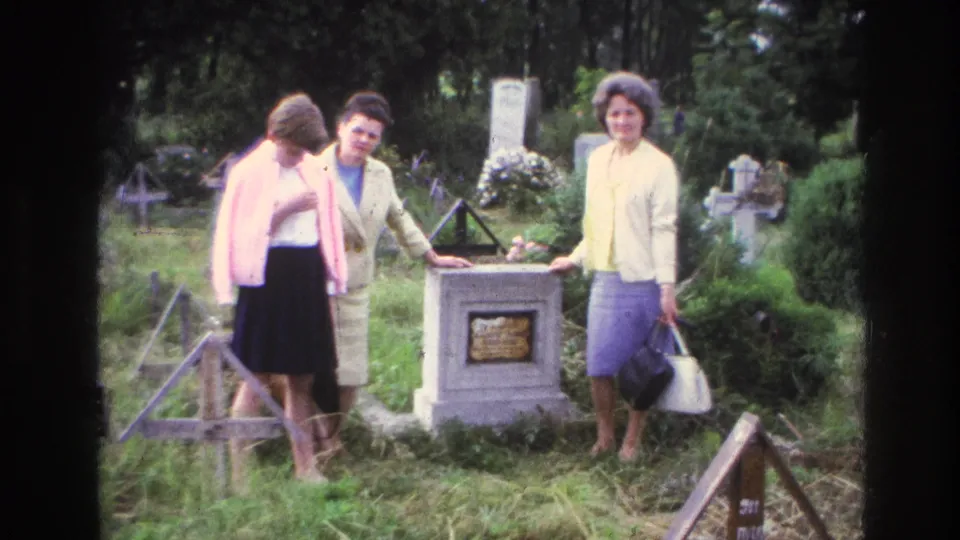 1967 France 8mm frame of three women in period clothing standing beside a stone memorial grave in a leafy cemetery.
