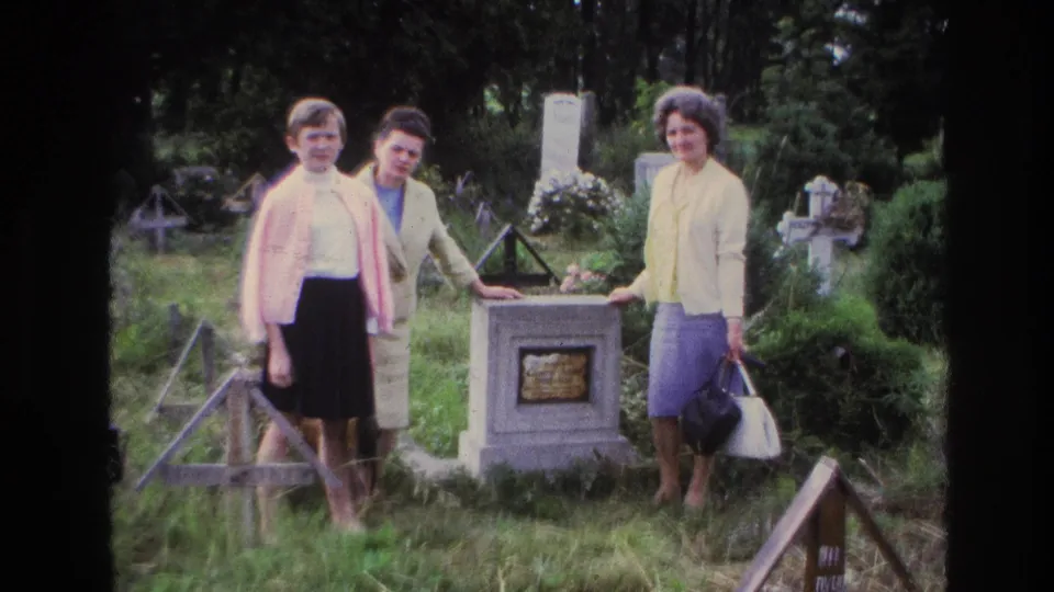 1967 France 8mm frame showing three women facing the camera beside a stone grave marker in a green cemetery setting.