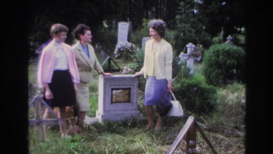 Three women in 1960s attire stand near a grave marker in a French cemetery, with crosses and greenery around them.