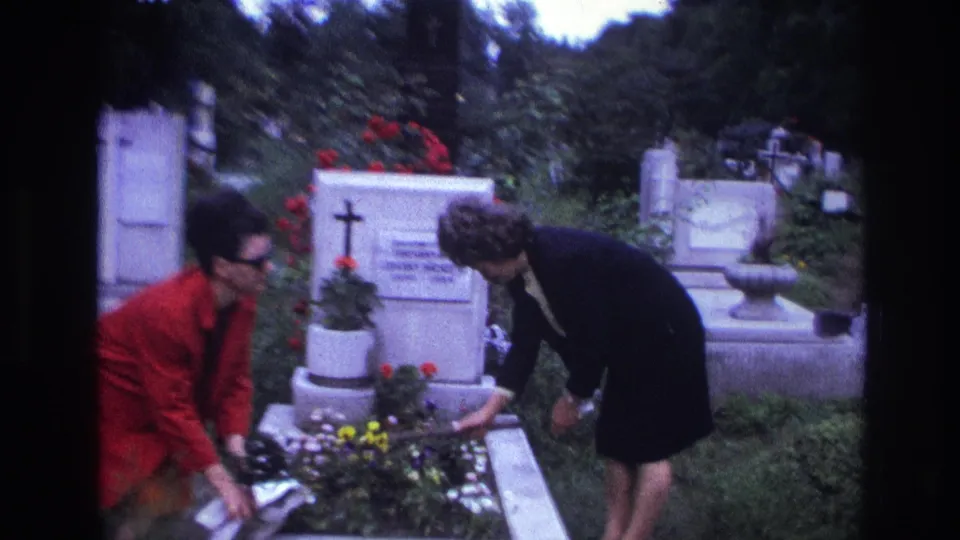 In a French cemetery, two women lean over a grave bed and adjust floral tributes beside a white stone marker.