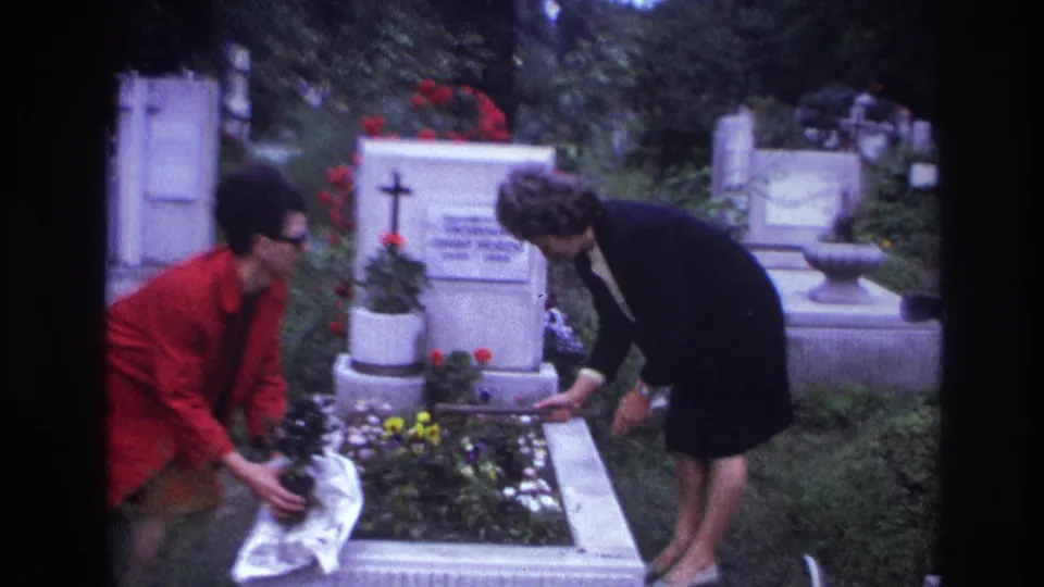 Two women continue tending a flower-covered grave in a French cemetery, framed by stone monuments and greenery.