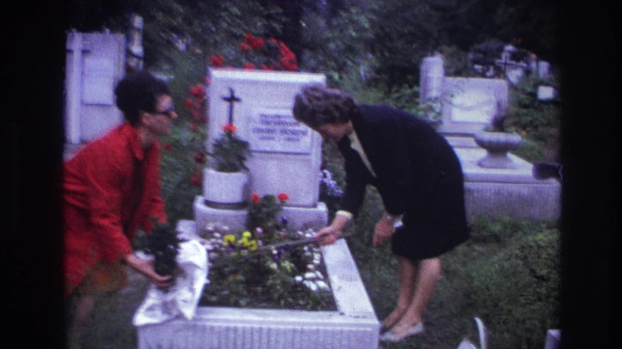 In 1967 France, two women appear to decorate a white grave plot with flowers in a green cemetery setting.