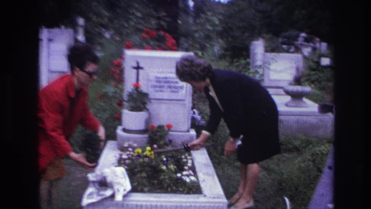 Two women in a French cemetery tend a grave with potted plants and flowers, with cross-shaped markers visible behind them.