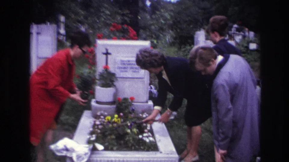 Three visitors stand close to a decorated cemetery grave as one woman adjusts flowers within the white border.