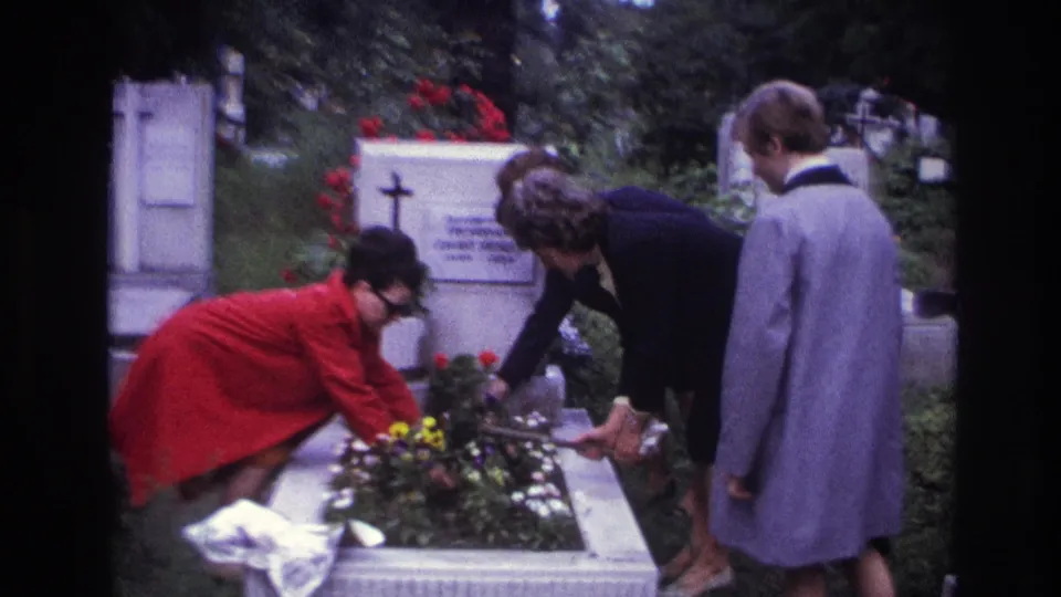 Home-movie frame from France in 1967 showing adults leaning over a grave bed with flowers and a white headstone behind them.