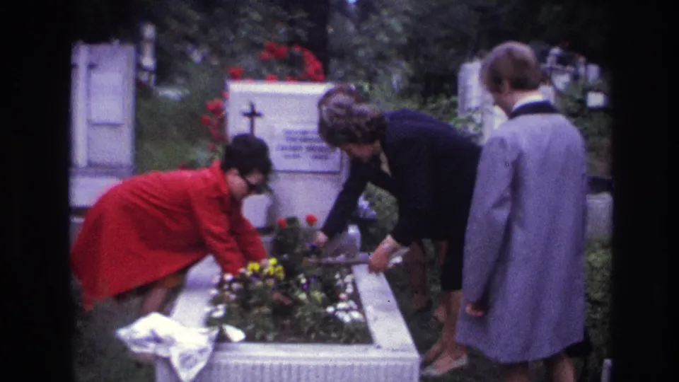 1967 French 8mm cemetery scene with adults gathered around a grave, continuing to set flowers below a white headstone.