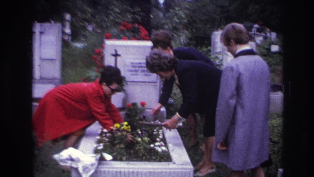 In France 1967 home-movie footage, three women in coats tend flowers on a white cemetery grave surrounded by headstones.