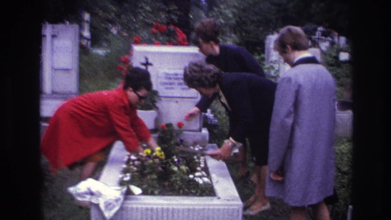 Three women in period coats carefully place flowers and adjust a plant on a grave in a French cemetery.