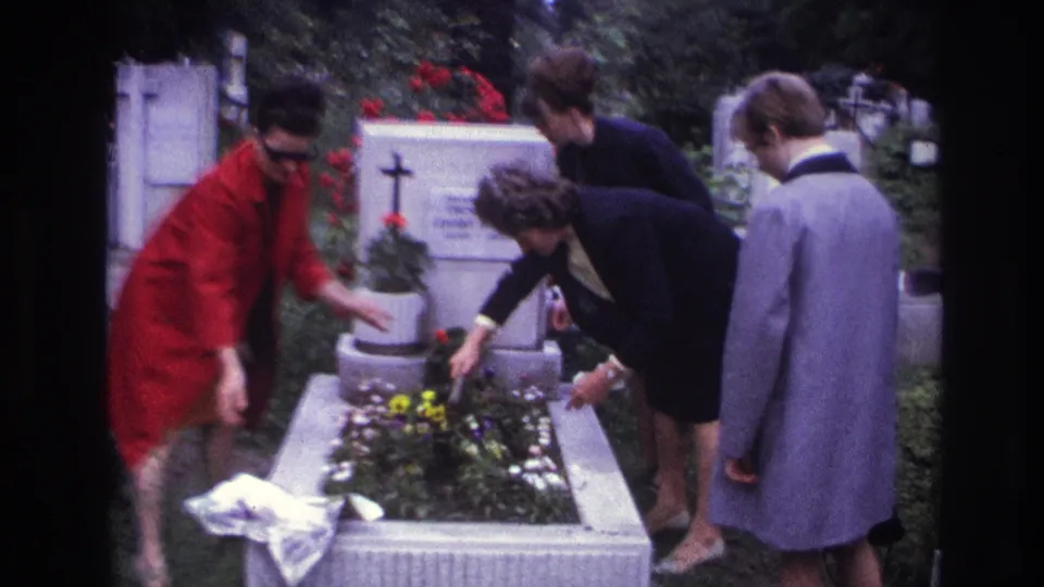 A woman in a red coat steps back while another bends over the grave flowers, with a third woman watching nearby.