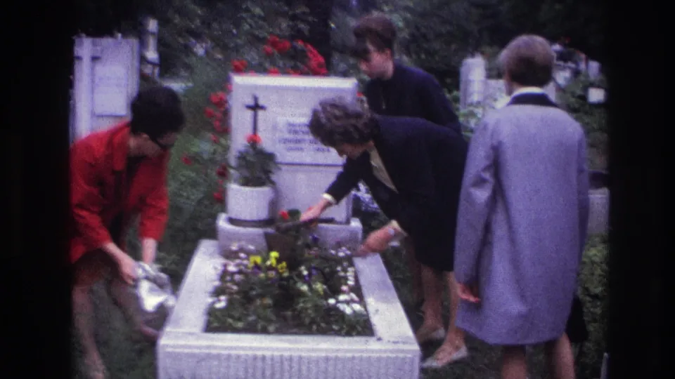 The cemetery visit ends with two women near the grave and a woman in red moving beside the white stone border.