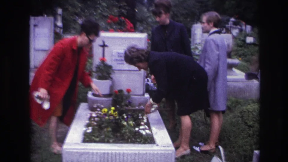 At a French cemetery grave, one woman reaches toward the planter while others observe near the white headstone and flowers.